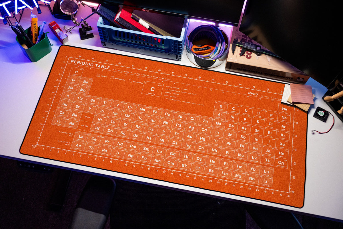 Periodic Table Pad desk mat in Orange / White, shown on a styled desk setup
