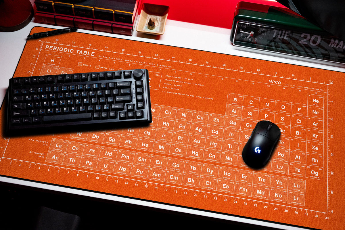 Periodic Table Pad desk mat in Orange / White, shown on a styled desk setup