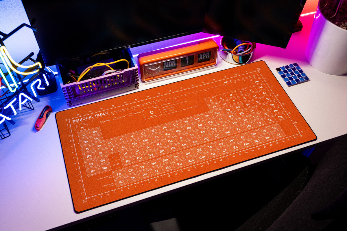 Periodic Table Pad desk mat in Orange / White, shown on a styled desk setup